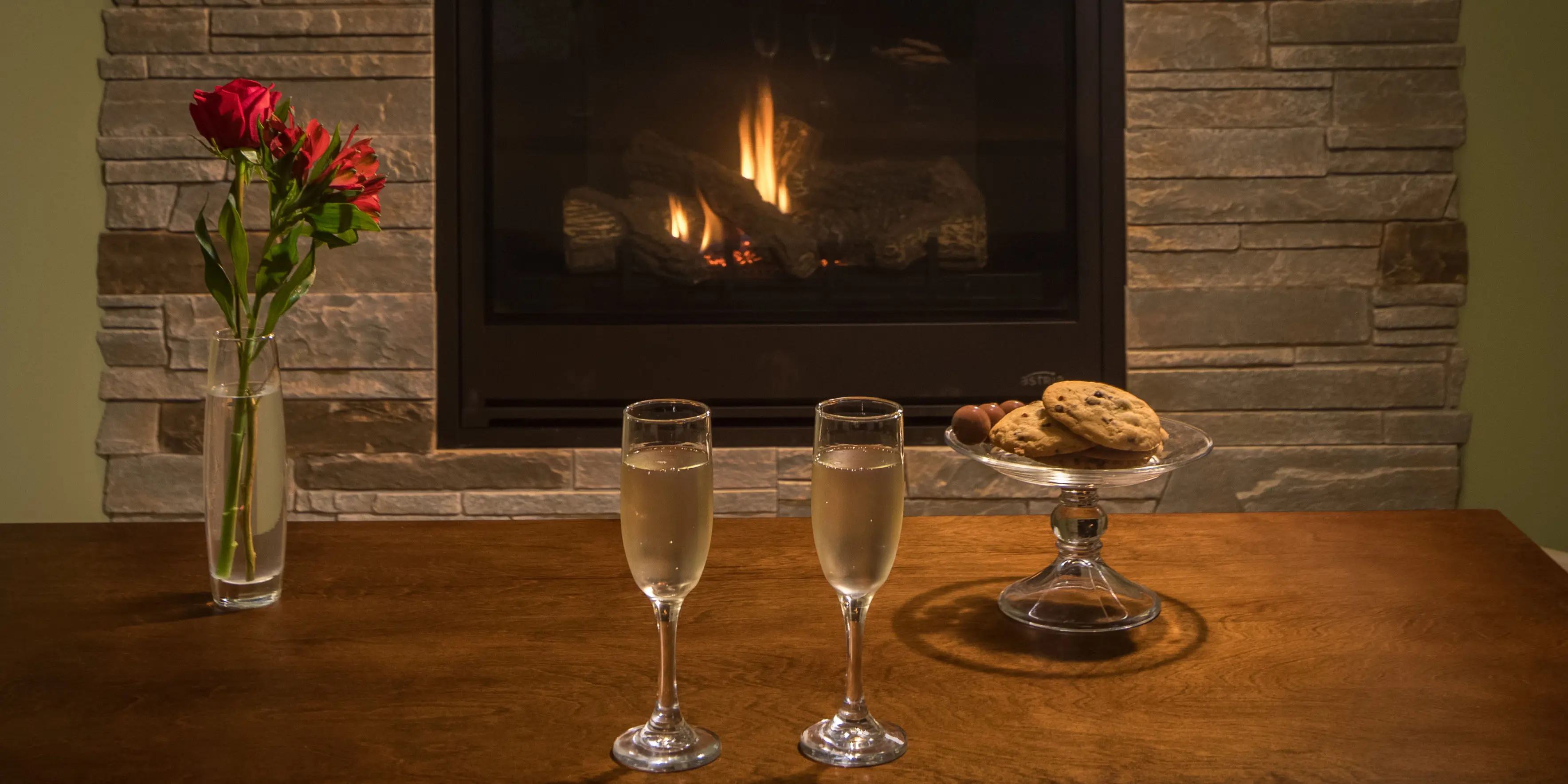 two glasses of champagne, cookies and flowers in front of stone fireplace