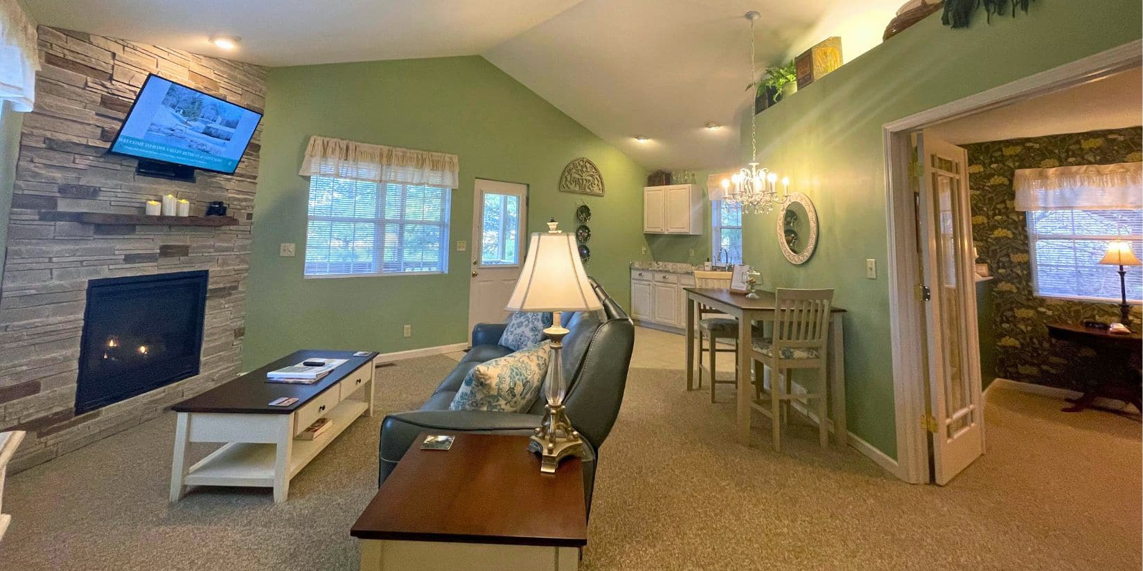 the somerset cottage with green wall living room, grey couch, stone fireplace, kitchen and bedroom in background